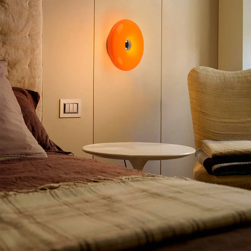 Orange, donutförmige wiederaufladbare Wandlampe an einer hellbeigen Wand über einem Bett.
