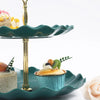 A two-tier serving stand in teal ceramic with a gold pedestal holding desserts and fruit.