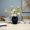 A black modern round metal decorative vase filled with light pink and green floral branches on a wooden table.