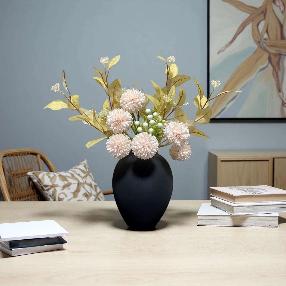 A black modern round metal decorative vase filled with light pink and green floral branches on a wooden table.
