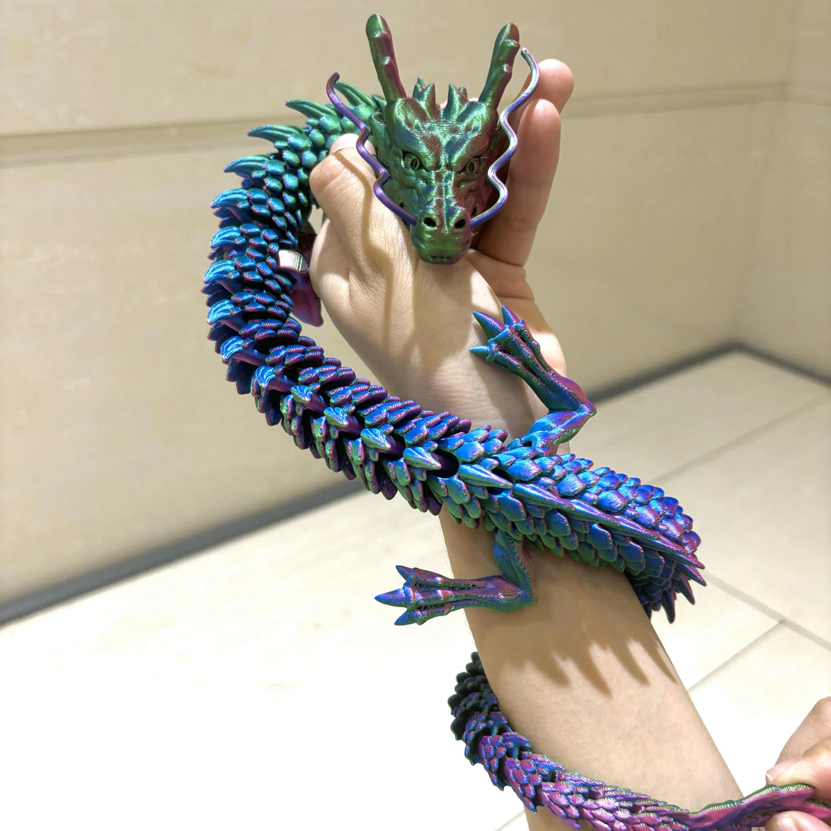 A hand holds a vibrant, articulated 3D printed dragon figurine with iridescent green and blue scales.