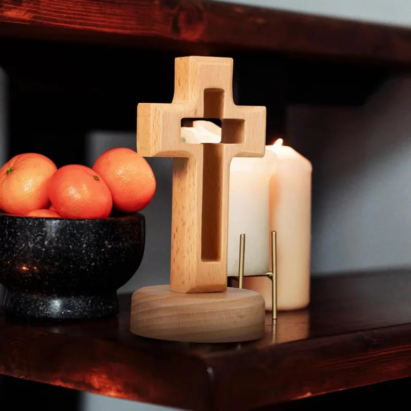 A wooden crucifix wall ornament with a cross carved from light wood sits next to a lit candle on a dark shelf.