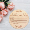 A round wooden baby name announcement sign with a leafy border and lines for name, date, weight, and length is placed on a wh