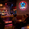 A modern coffee bar features a neon sign shaped like a cup with the word Coffee glowing brightly on a textured wall.