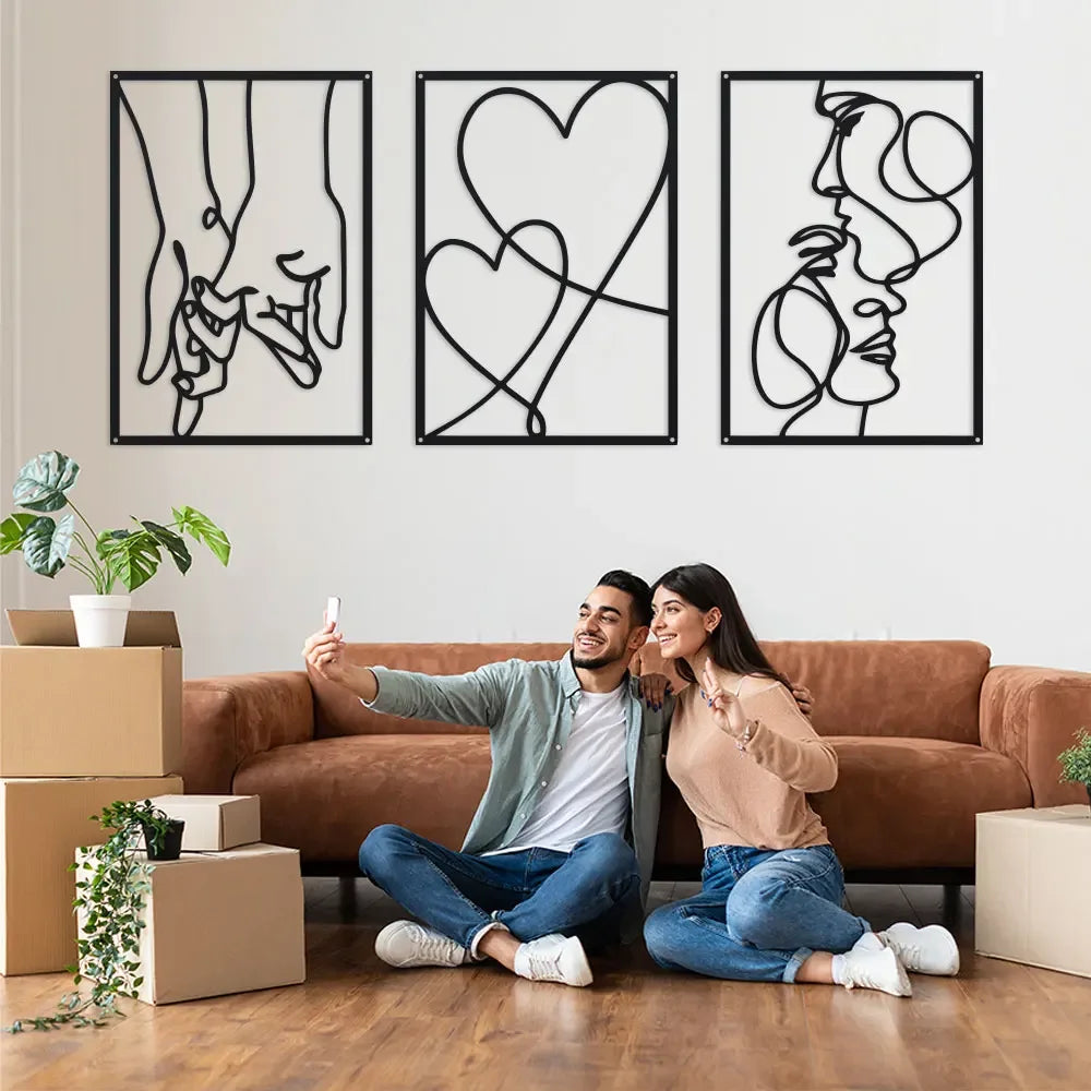 A couple sitting on a couch in front of three framed black line art prints depicting holding hands, love, and kissing motifs.