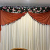 A white backdrop curtain decorated with floral garlands and draped in orange fabric for an event.