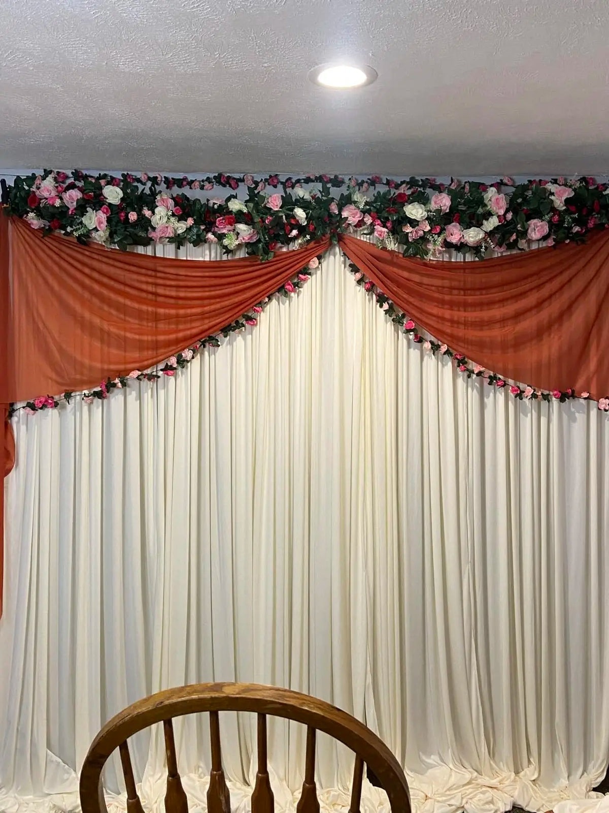 A white backdrop curtain decorated with floral garlands and draped in orange fabric for an event.