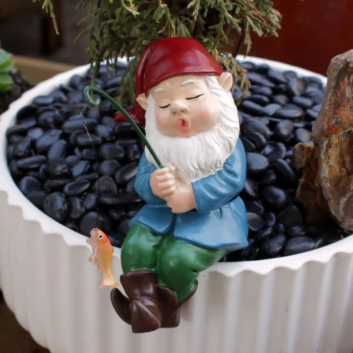 Keramiks statue eines bärtigen Gnomens mit rotem Hut und blauer Kleidung, der mit einer grünen Angelrute in eine Schale mit s