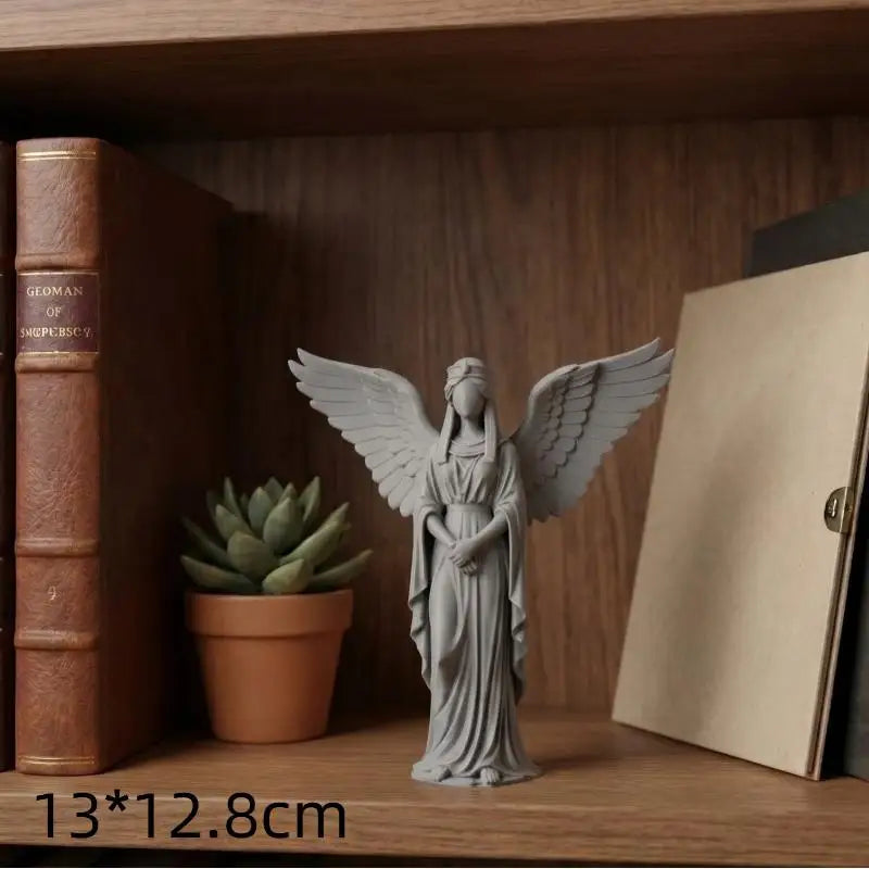 A gray praying angel figurine with wings is displayed on a wooden shelf next to a book.