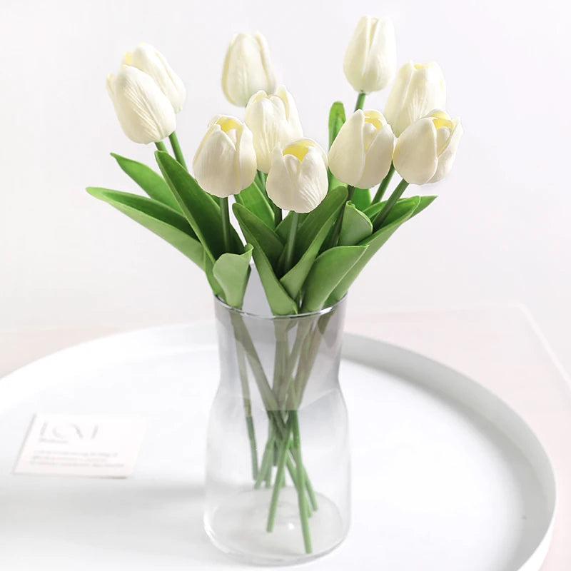 A bouquet of white artificial tulip flowers in a glass vase on a white plate.