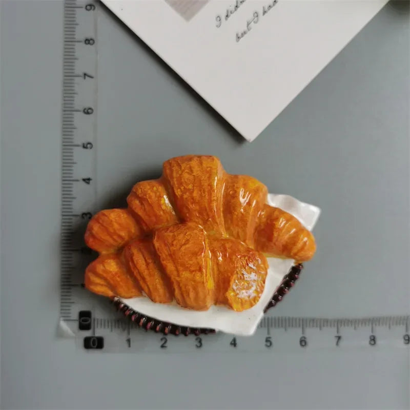 A golden croissant is shown on white paper, positioned next to a ruler and a piece of paper.