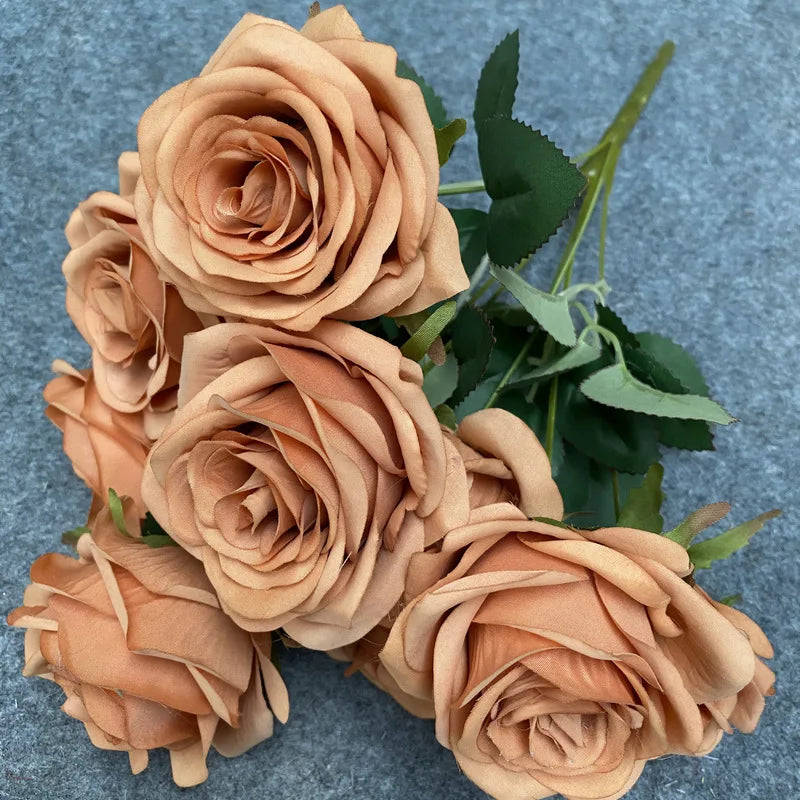 A bouquet of nine realistic silk roses in a soft peach or tan color against a gray background.