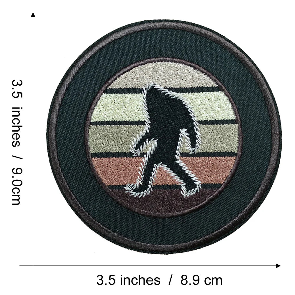 A round, dark brown embroidered patch featuring a silhouette of Bigfoot in white and gray fur against a beige background.