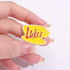 A hand holds a yellow enamel pin shaped like a retro television, featuring the text Lukes.