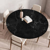 A round black marble print tablecloth is set on a wooden table with white chairs and a vase.