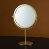 A round, gold-framed makeup mirror with a reflective surface mounted on a slender stand.