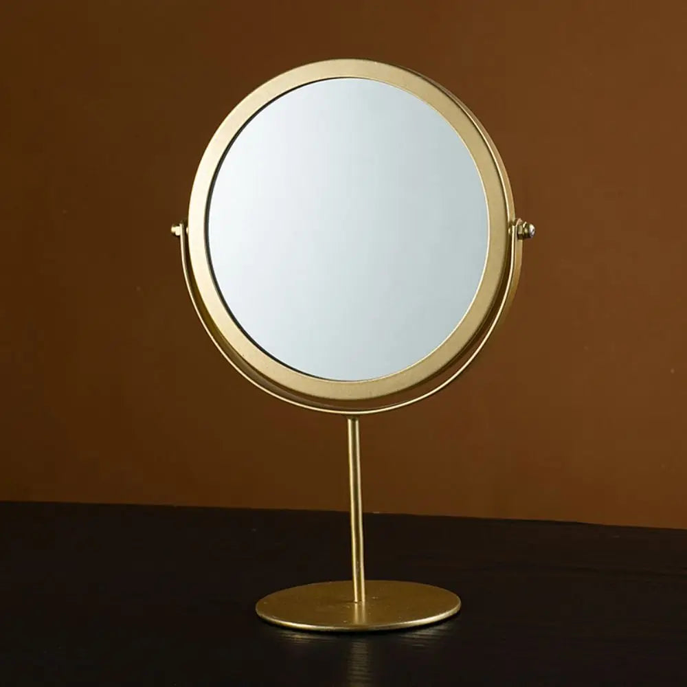 A round, gold-framed makeup mirror with a reflective surface mounted on a slender stand.