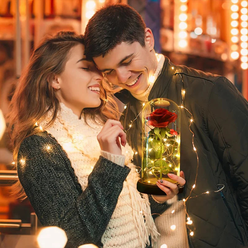 A couple is smiling while holding a glass dome containing an eternal rose surrounded by twinkling fairy lights.