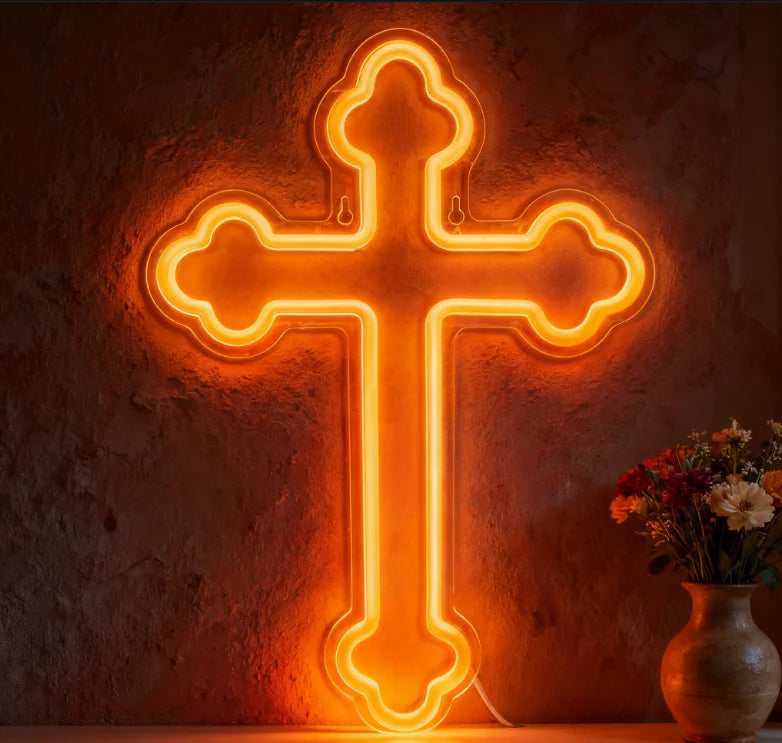 A glowing orange neon cross wall sign mounted on a textured stone wall next to a vase of flowers.