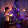 A young boy stands next to a brightly lit Christmas tree decorated with colorful lights and ornaments.