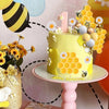 A yellow birthday cake decorated with honeycomb shapes, a number one topper, and small bee figures on a pink stand.