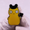A yellow enamel pin badge shaped like a funny burger with black hair and eyes, held against a white background.