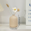 A minimalist ceramic vase with a speckled beige finish holds dried floral elements on a white shelf.