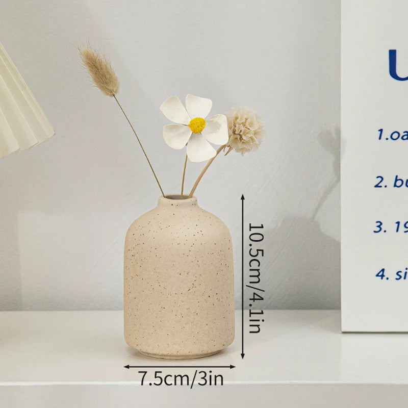 A minimalist ceramic vase with a speckled beige finish holds dried floral elements on a white shelf.