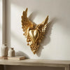 Gold ornate winged heart wall decor mounted on a white wall above a wooden shelf.