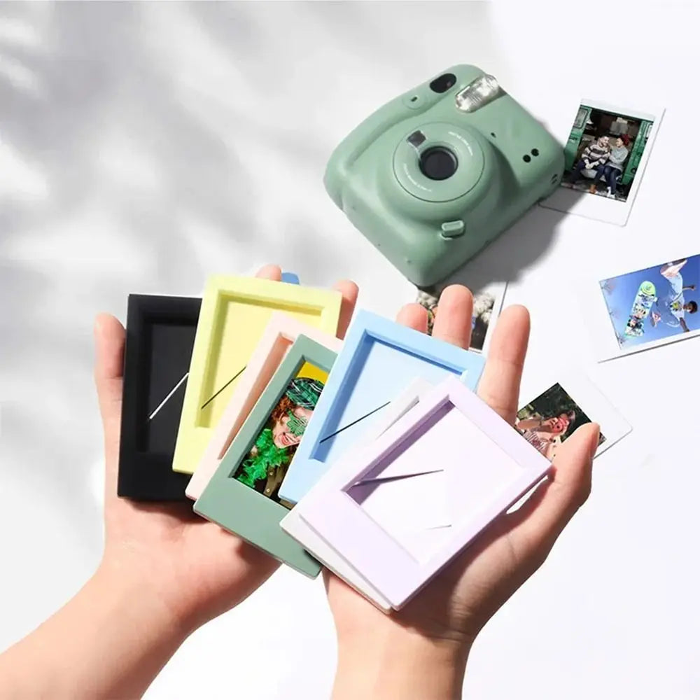 A hand holds a green mini film frame displaying an instant photo, with several other colorful frames nearby.