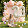A woman sits in a grassy field surrounded by pink and gold balloon arches and balloons, with large foil number balloons nearb