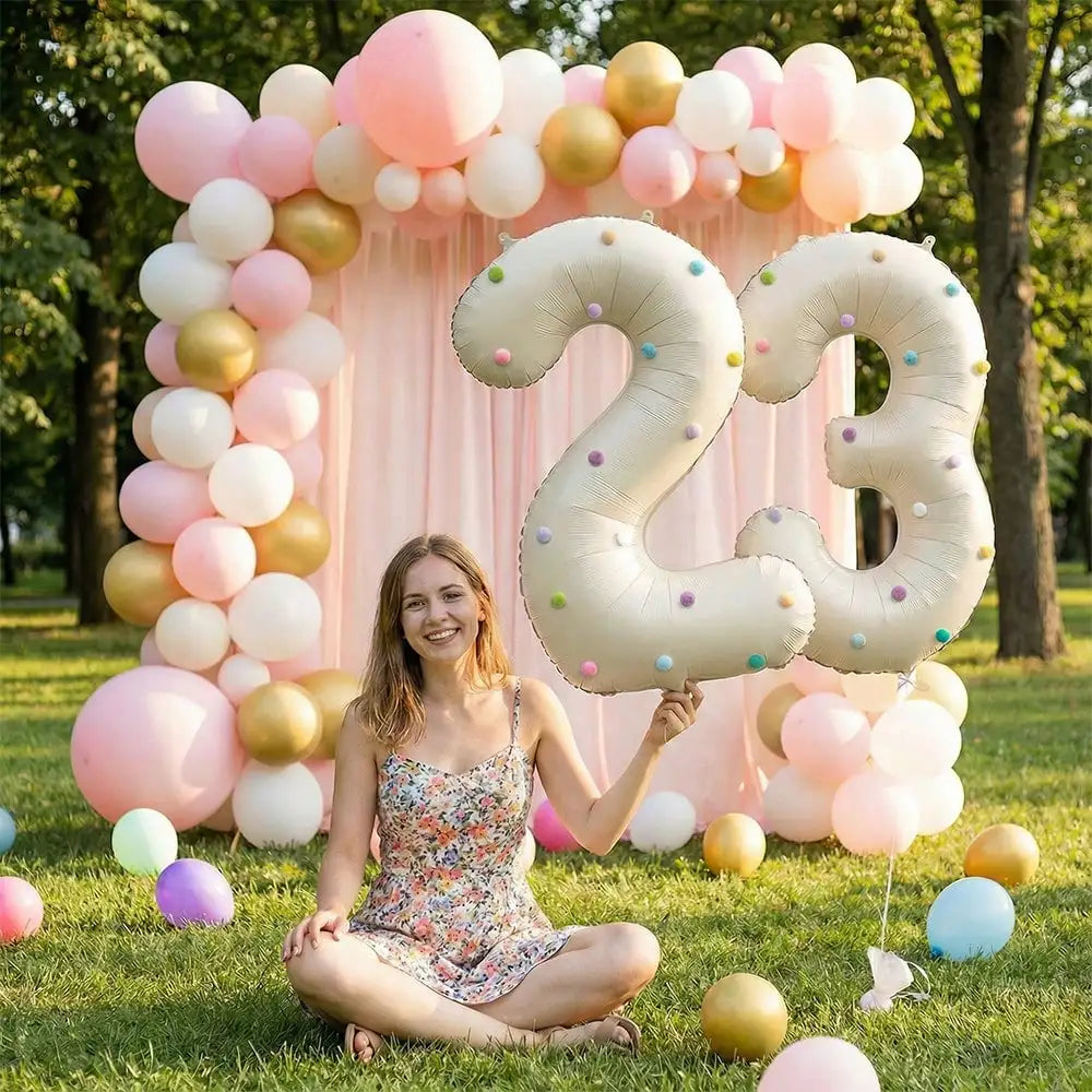 A woman sits in a grassy field surrounded by pink and gold balloon arches and balloons, with large foil number balloons nearb