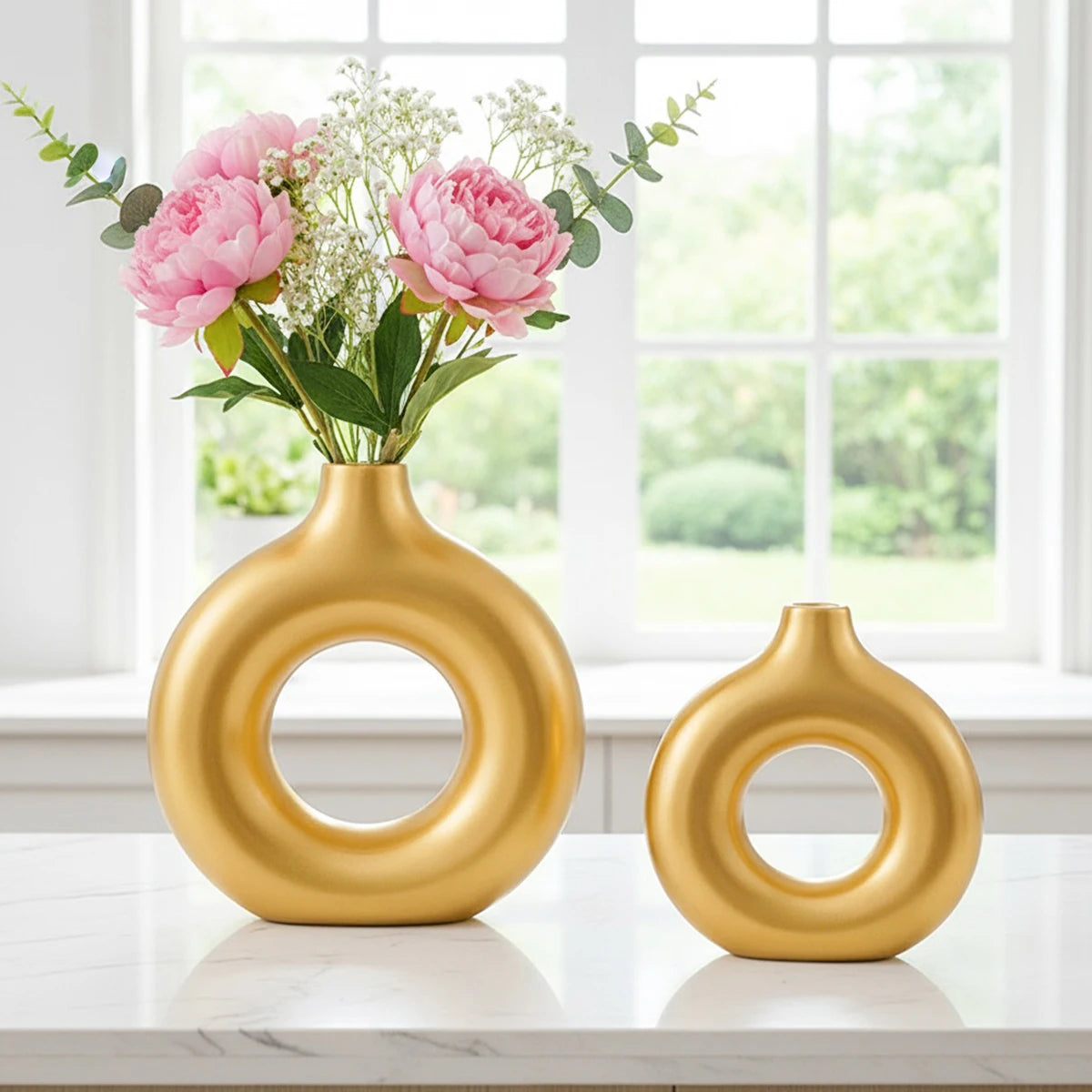 A golden, ring-shaped resin vase filled with pink and white flowers sits on a white marble surface near a window.