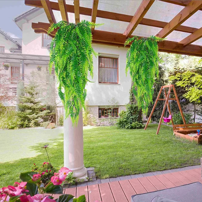 A long green artificial ivy vine garland hangs from a white column under a wooden pergola in a garden setting.