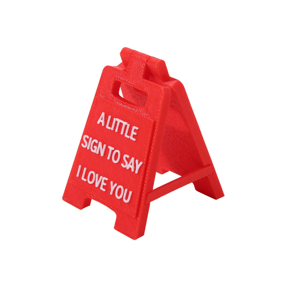 A red 3D printed letter sign ornament shaped like a house with the text A LITTLE SIGN TO SAY I LOVE YOU engraved on it.