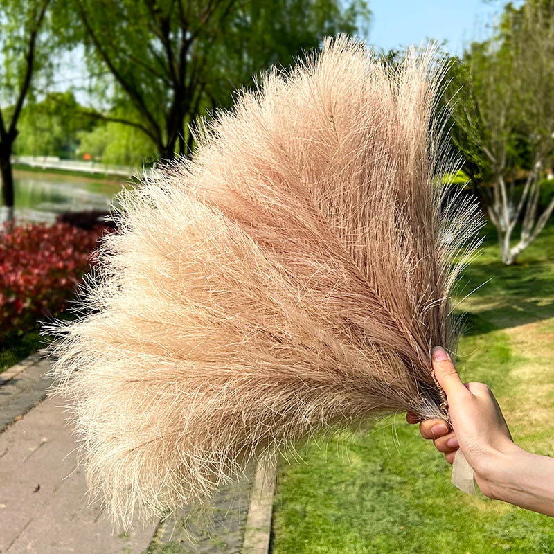 Una mano sostiene un gran ramo artificial de pampas de color beige claro para decoración del hogar.