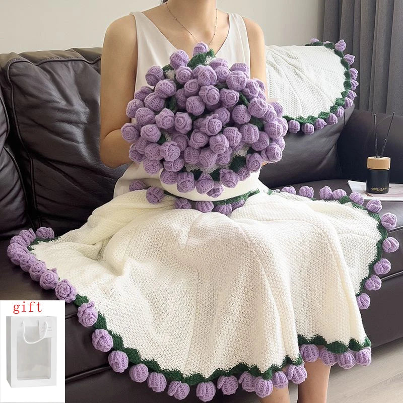 A woman is wearing a white knitted blanket with purple floral trim, sitting on a dark sofa.