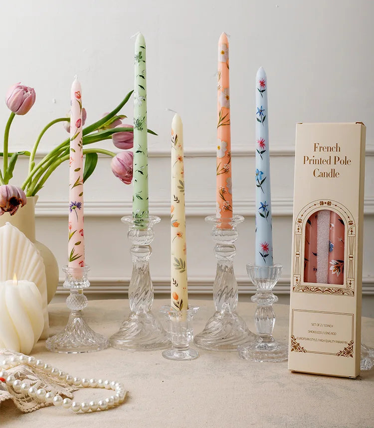 A collection of decorative printed long stick candles in various colors and floral patterns displayed on a light surface.