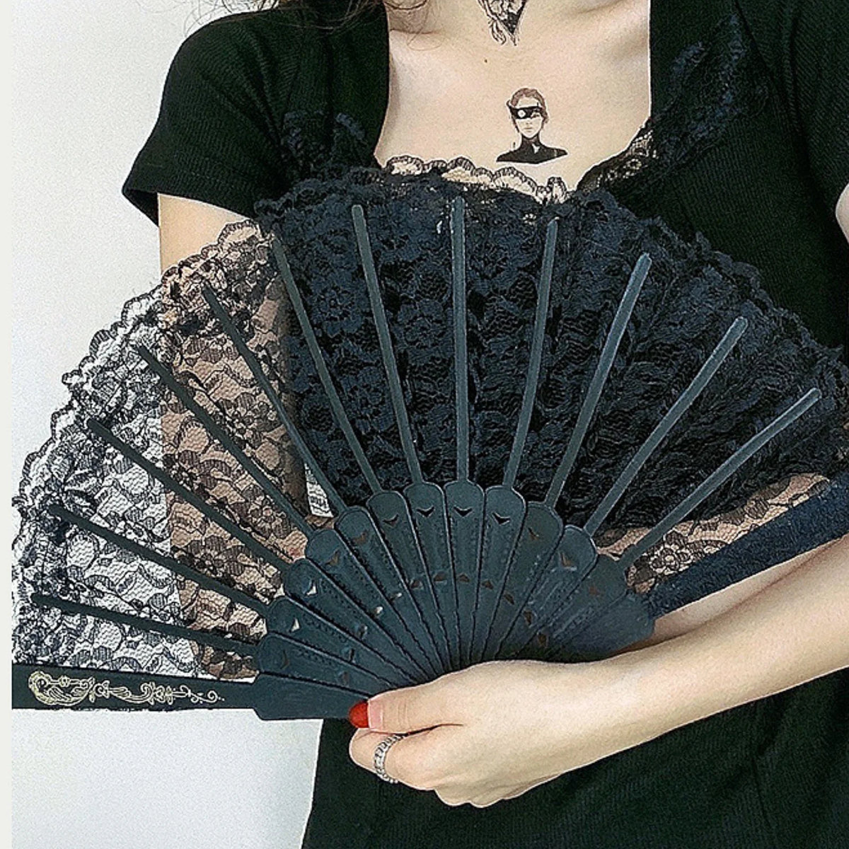 Una mujer sostiene un abanico plegable de encaje oscuro con acentos en negro y beige contra un top negro.