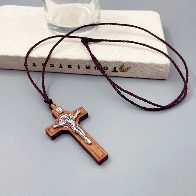 A wooden cross necklace featuring a crucifix pendant and a dark brown waxed rope cord, resting on a light-colored surface.