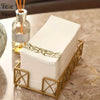 A stack of white paper dinner napkins with rose gold trim is displayed in a gold-colored metal holder.