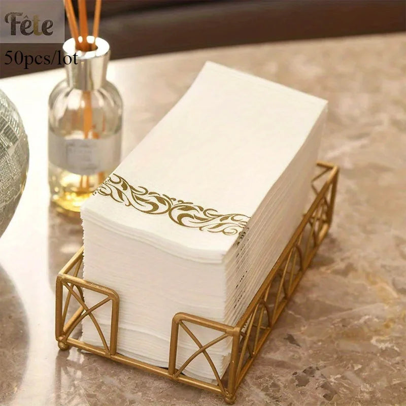 A stack of white paper dinner napkins with rose gold trim is displayed in a gold-colored metal holder.