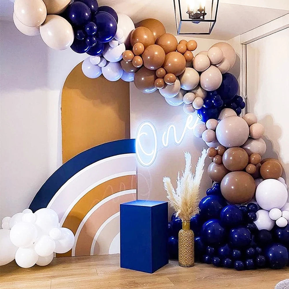 A balloon arch decoration featuring assorted latex balloons in shades of white, blue, and brown against a light wall.