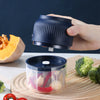 A hand holds a dark blue electric mini food chopper with a clear container filled with vegetables on a wooden cutting board.