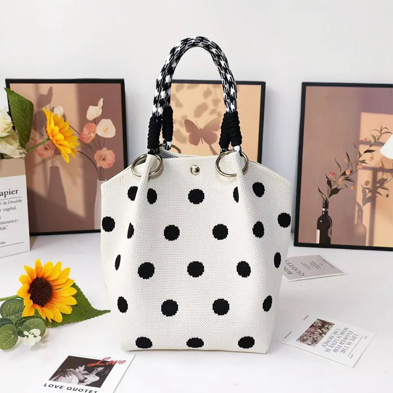 A white canvas tote bag with black polka dots and a woven black and white handle, displayed next to floral arrangements.