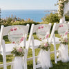 A row of white folding chairs decorated with pink floral thank you hand fans for an outdoor event.