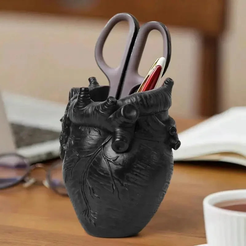 A black heart-shaped resin pen organizer with a silver-colored pen inside, resting on a wooden desk.