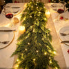 A realistic cedar garland with flexible branch design decorated with warm lights and candles on a dining table.