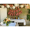 A festive birthday decoration featuring pink and gold foil letter balloons arranged in a garland against a rustic wall.