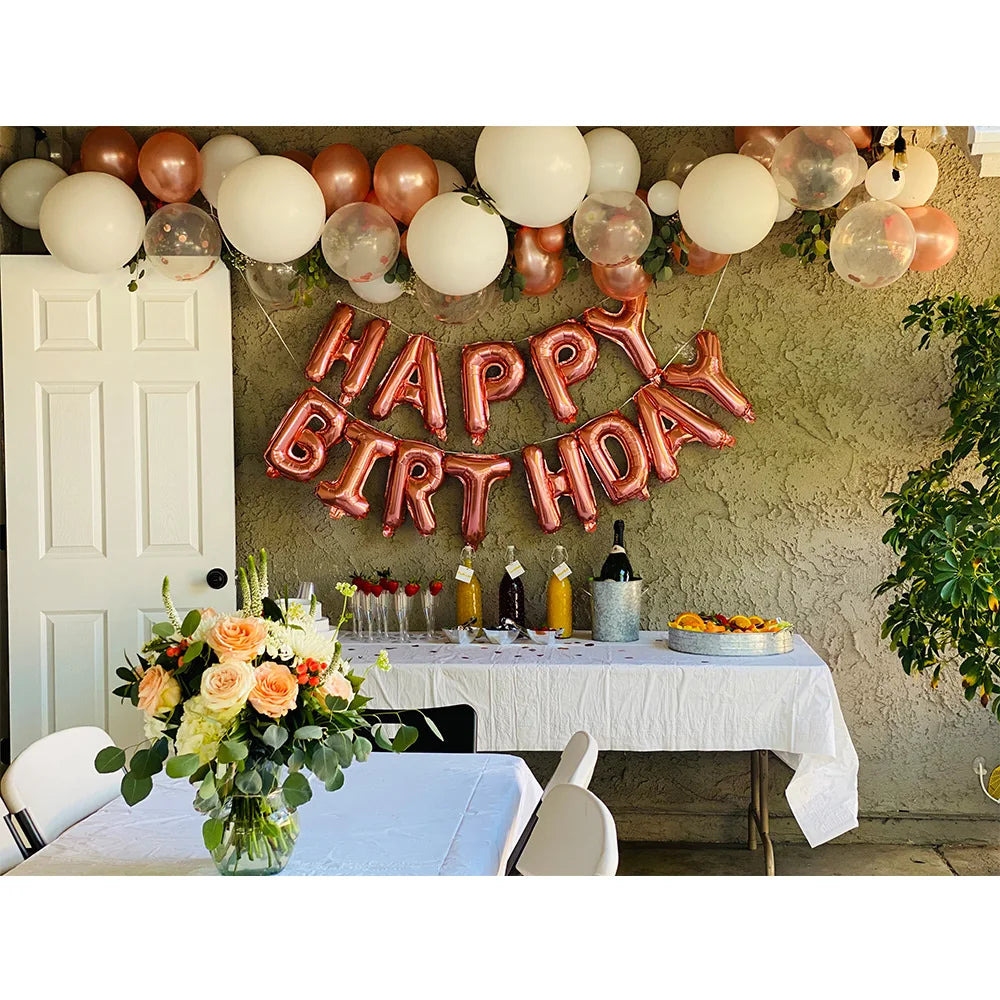 A festive birthday decoration featuring pink and gold foil letter balloons arranged in a garland against a rustic wall.